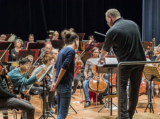 Orchester im Hintergrund, davor steht der Dirigent und eine Teilnehmer*in des Wettbewerbs